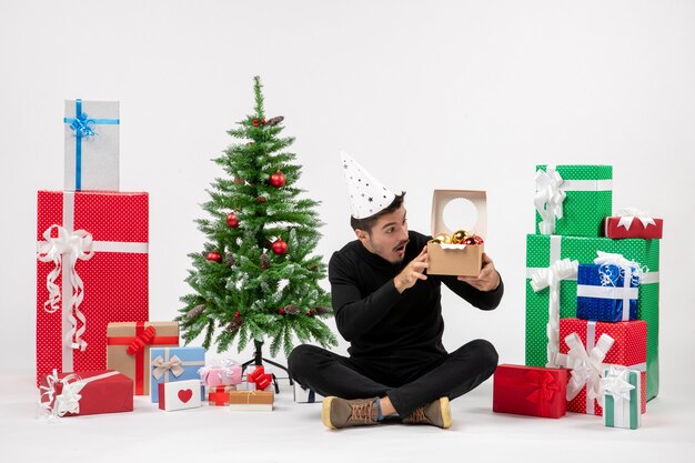 Widok z przodu młodego mężczyzny siedzącego wokół prezentów świątecznych trzymających zabawki drzewne na białej ścianie