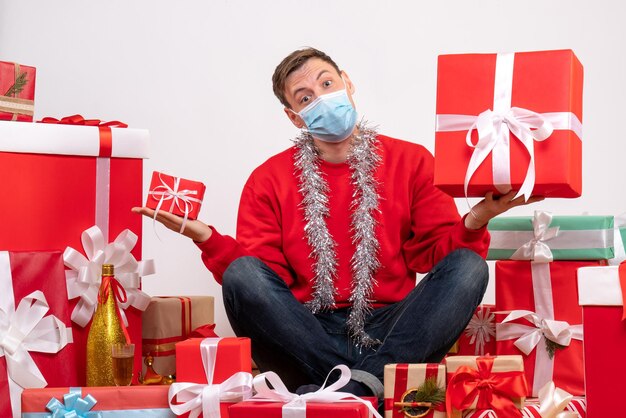 Widok z przodu młodego mężczyzny w sterylnej masce siedzącego wokół świątecznych prezentów na białej ścianie