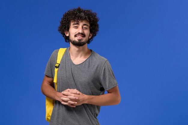 Widok z przodu młodego mężczyzny w szarym t-shircie ubrany w żółty plecak uśmiechnięty na niebieskiej ścianie