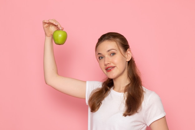 Widok z przodu młodej kobiety w białej koszulce trzymającej zielone jabłko na różowej ścianie