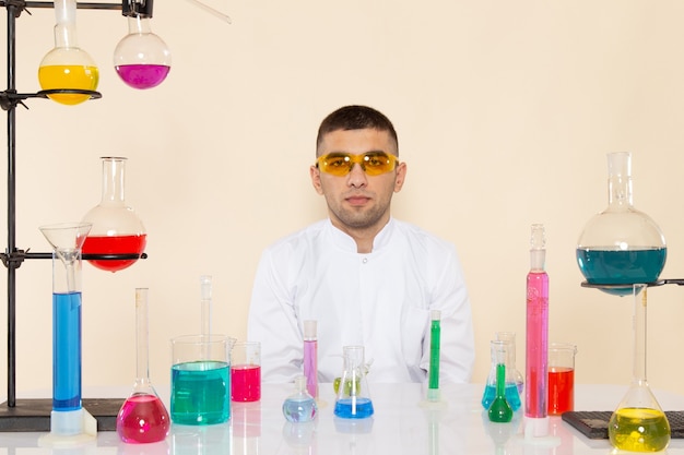 Widok z przodu młody chemik mężczyzna w białym specjalnym garniturze siedzi przed stołem z roztworami na kremowej ścianie laboratorium eksperyment naukowy chemii
