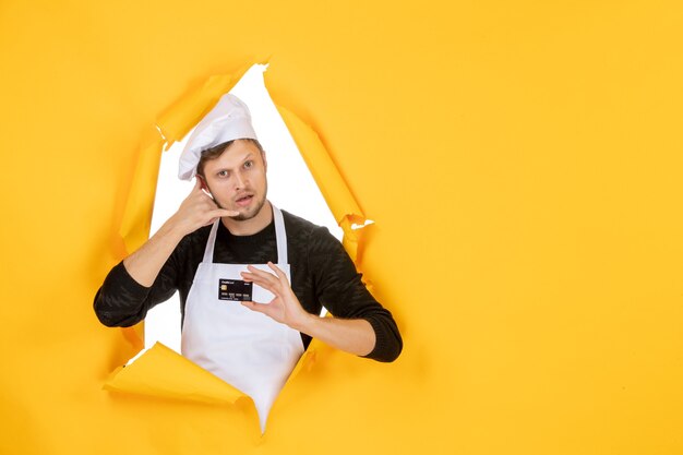 Widok z przodu młody kucharz mężczyzna w białej pelerynie trzymający czarną kartę bankową na żółtym tle model biały kolor kuchnia praca człowiek jedzenie pieniądze