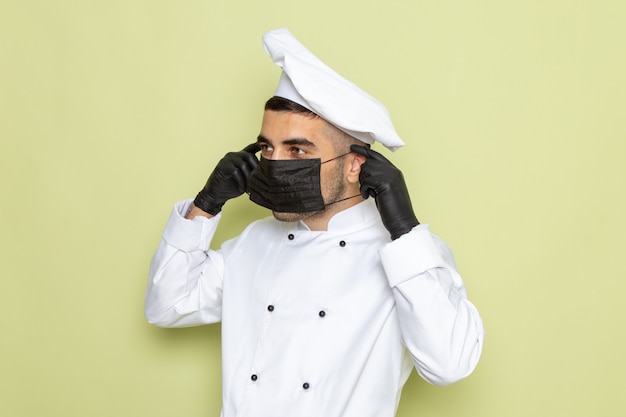 Bezpłatne zdjęcie widok z przodu młody mężczyzna kucharz w białym garniturze kucharza w ciemnych rękawiczkach i masce na zielonym mundurze kuchni męskiej pracy