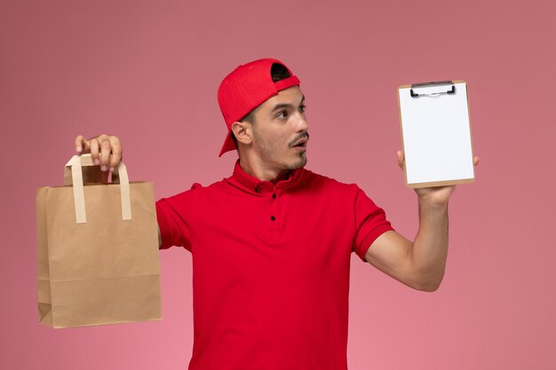 Widok z przodu młody mężczyzna kurier w czerwonej pelerynie mundurze, trzymając pakiet żywności i notatnik na różowym tle.