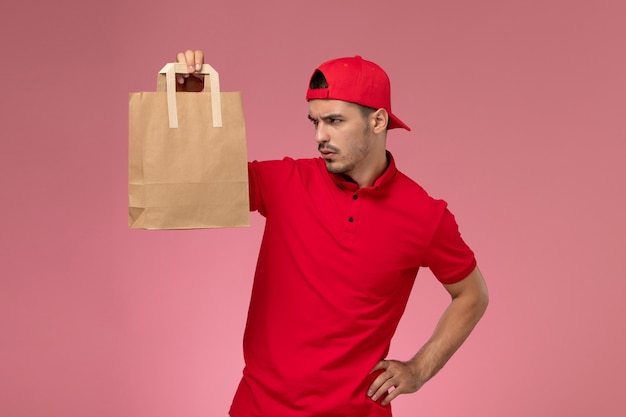 Bezpłatne zdjęcie widok z przodu młody mężczyzna kurier w czerwonej pelerynie mundurze trzymając papierowy pakiet żywności na różowym biurku.