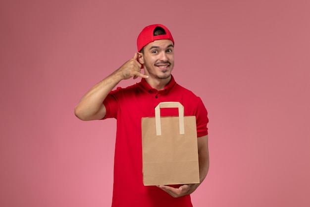 Widok z przodu młody mężczyzna kurier w czerwonej pelerynie mundurze trzymając papierowy pakiet żywności na różowym tle.
