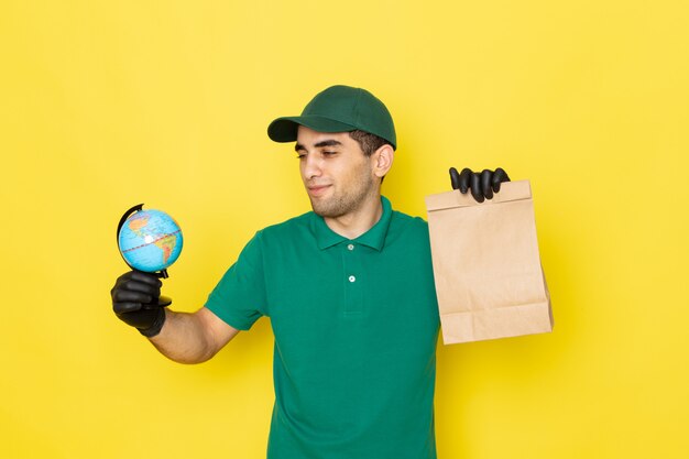 Widok z przodu młody mężczyzna kurier w zielonej koszuli zielonej czapce, trzymając pakiet dostawy i kula ziemska na żółto