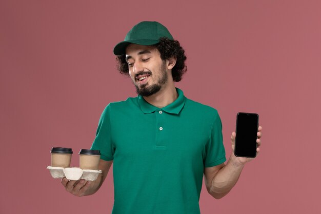 Widok z przodu młody mężczyzna kurier w zielonym mundurze i pelerynie, trzymając filiżanki kawy dostawy i telefon na różowym tle