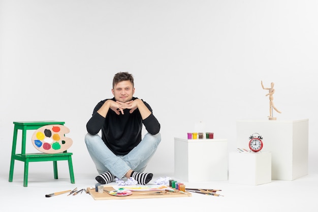 Widok z przodu młody mężczyzna siedzący wokół farb i frędzli do rysowania na białej ścianie rysuj kolorowe zdjęcia artysta maluje malarstwo artystyczne