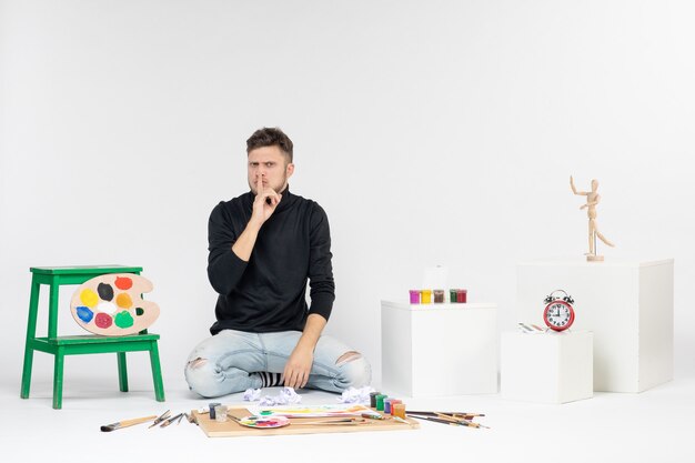 Widok z przodu młody mężczyzna siedzący wokół farb i frędzli do rysowania na białych farbach ściennych artysta rysuje malarstwo artystyczne