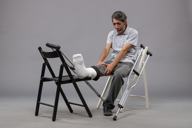 Bezpłatne zdjęcie widok z przodu młody mężczyzna siedzący ze złamaną stopą i kulami krzyczący z bólu na szarej ścianie ból stopy wypadek noga złamany skręt