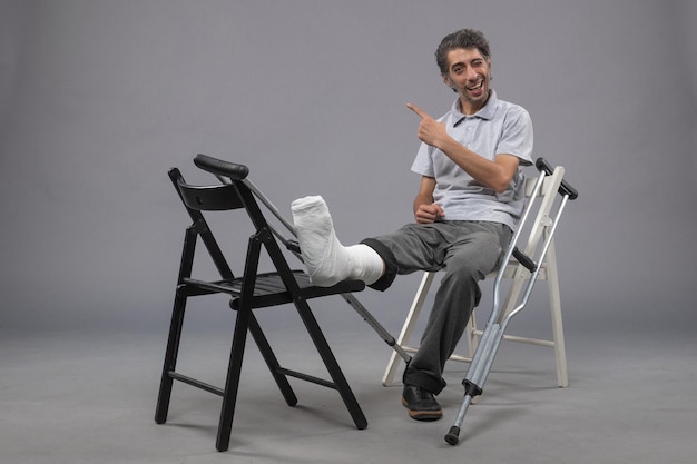 Bezpłatne zdjęcie widok z przodu młody mężczyzna siedzący ze złamaną stopą i kulami na szarej ścianie ból wypadek złamany skręt nogi noga