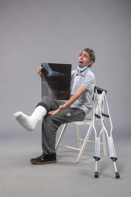 Widok z przodu młody mężczyzna siedzi ze złamaną stopą i trzyma prześwietlenie na szarej ścianie ból nogi wypadek skręcenie stopy mężczyzna