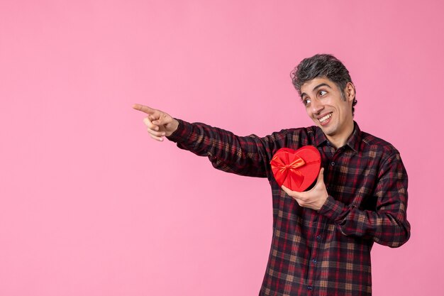 Widok z przodu młody mężczyzna trzyma prezent w kształcie czerwonego serca na różowej ścianie