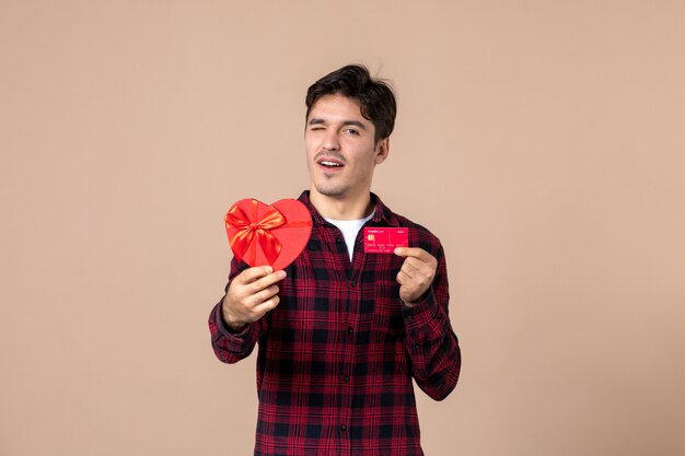 Widok z przodu młody mężczyzna trzymający prezent w kształcie serca i kartę bankową na brązowej ścianie