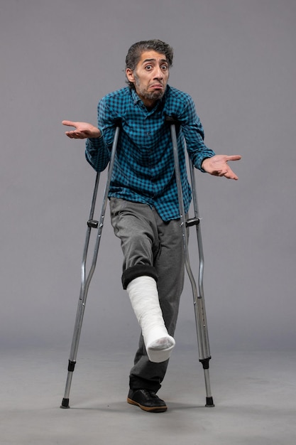Bezpłatne zdjęcie widok z przodu młody mężczyzna używający kul z powodu złamanej stopy na szarej ścianie noga wypadek wyłącz złamany uszkodzony