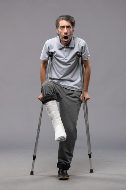Widok z przodu młody mężczyzna używający kul z powodu złamanej stopy na szarej ścianie wyłącza wypadek złamanej stopy