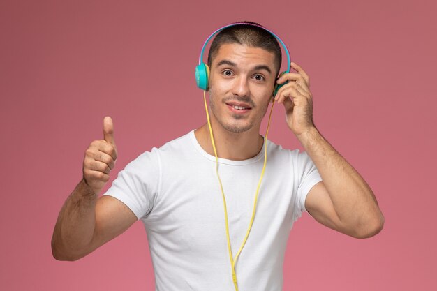 Widok z przodu młody mężczyzna w białej koszulce, słuchający muzyki przez słuchawki na jasnoróżowym biurku