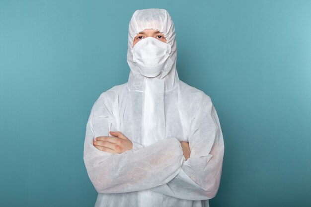 Widok z przodu młody mężczyzna w białym specjalnym garniturze, ubrany w sterylną maskę ochronną, pozujący na niebieskiej ścianie, mężczyzna w kolorze niebezpiecznego wyposażenia specjalnego