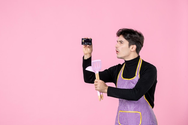 Widok z przodu młody mężczyzna w pelerynie trzymający kartę bankową i łyżki na różowym tle poziome jednolite praca gotowanie pracownik kolor kuchnia zawód szef pieniądze
