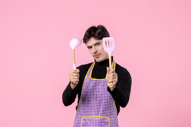 Widok z przodu młody mężczyzna w pelerynie trzymający łyżki na różowym tle zawód kolor posiłek gotowanie kuchnia pozioma praca jednolite ciasto