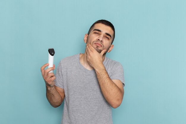 Widok z przodu młody mężczyzna w szarej koszulce trzymający elektryczną maszynkę do golenia na lodowoniebieskiej męskiej piance do golenia brody