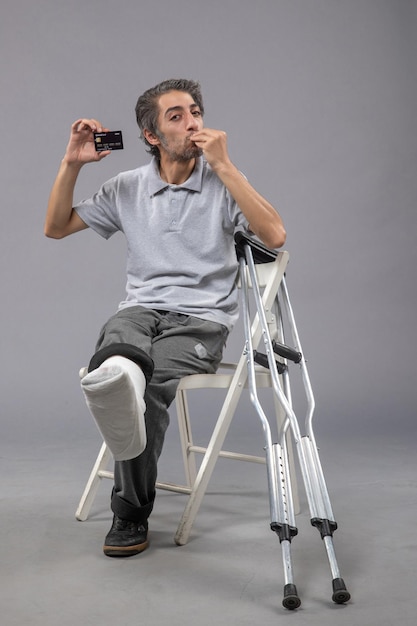 Bezpłatne zdjęcie widok z przodu młody mężczyzna ze złamaną stopą i bandażem trzymający czarną kartę bankową na szarej ścianie ludzki skręt męski wypadek ból stóp