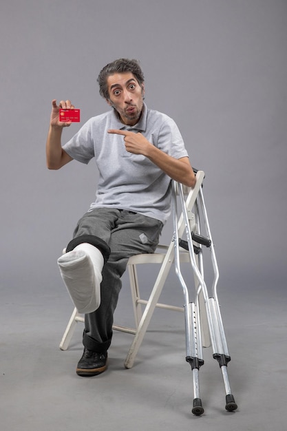Bezpłatne zdjęcie widok z przodu młody mężczyzna ze złamaną stopą i bandażem trzymający kartę bankową na szarej podłodze wypadek skręcenie nogi stopa ludzki ból mężczyzna