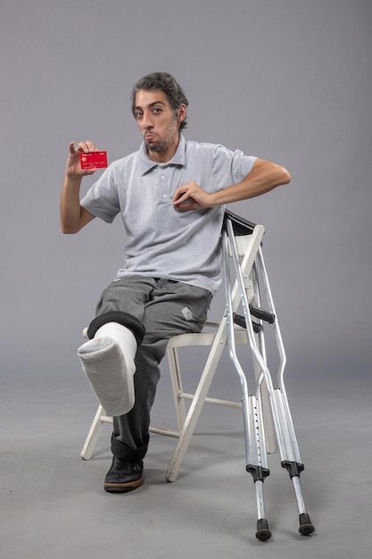 Widok z przodu młody mężczyzna ze złamaną stopą i bandażem trzymający kartę bankową na szarej ścianie męski wypadek skręca ból ludzkiej stopy