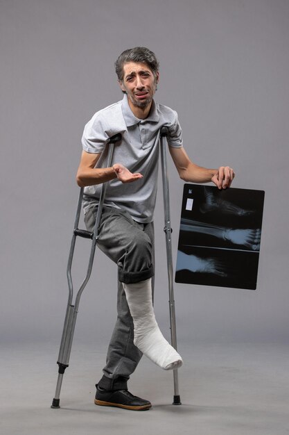 Widok z przodu młody mężczyzna ze złamaną stopą używający kul i trzymający prześwietlenie na szarej podłodze ból wyłącza złamaną stopę po wypadku