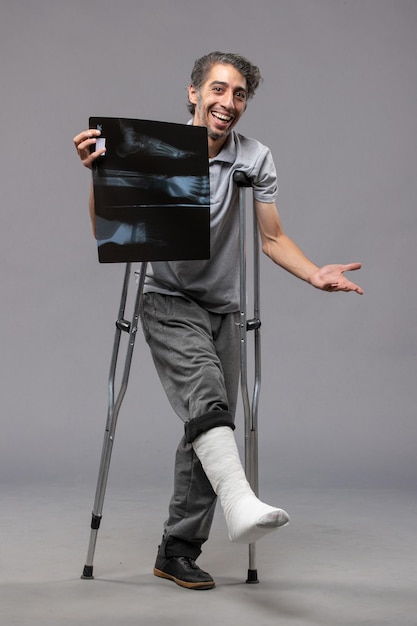 Widok z przodu młody mężczyzna ze złamaną stopą używający kul i trzymający prześwietlenie na szarym biurku wyłącza złamany ból w razie wypadku skręcenie stopy