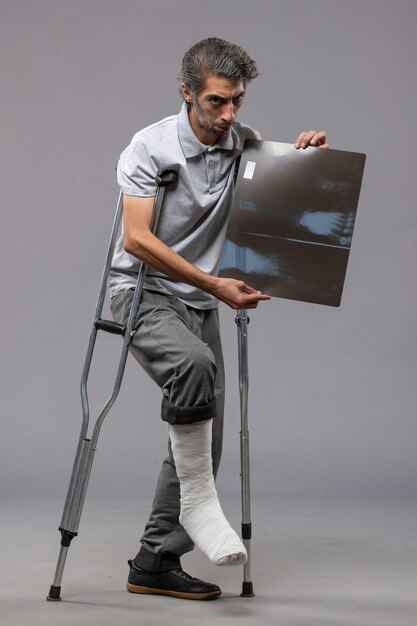 Widok z przodu młody mężczyzna ze złamaną stopą za pomocą kul i trzymający prześwietlenie na szarej ścianie ból wyłącza skręt złamanej stopy powypadkowej