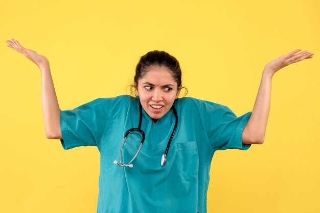 Widok z przodu mylić kobieta lekarz ze stetoskopem, otwierając ręce, stojąc na żółtym tle