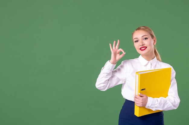 Widok z przodu nauczycielka pozuje z żółtymi plikami na zielonym tle szkolna książka lekcyjna praca student mundurek uniwersytecki studium