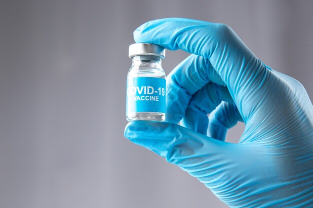 Widok z przodu niebieskiej rękawiczki noszącej dłoń trzymającą zamkniętą ampułkę ze szczepionką przeciw covid na szarym tle fali