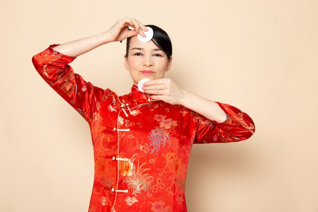 Widok z przodu piękna japońska gejsza w tradycyjnej czerwonej japońskiej sukience z paskami do włosów, pozowanie, trzymając trochę białej bawełny elegancki uśmiechnięty na ceremonii kremowej tle japonia