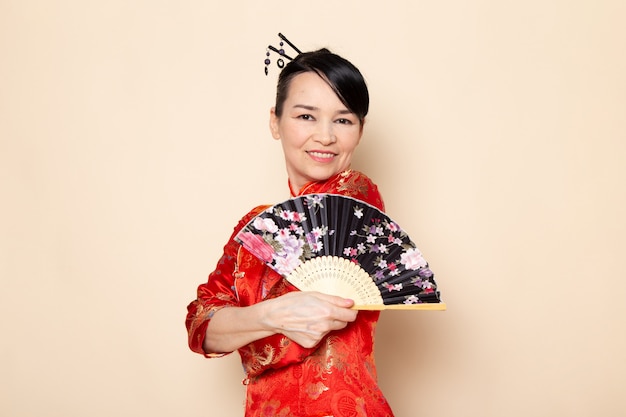 Widok Z Przodu Piękna Japońska Gejsza W Tradycyjnej Czerwonej Japońskiej Sukni Z Włosami, Stawiając Składane Wachlarz Elegancki Uśmiechnięty Na Ceremonii Kremowej Tle Japonia