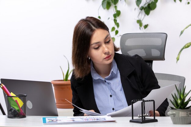 Widok z przodu piękna młoda bizneswoman w czarnej kurtce i niebieskiej koszuli pracującej z laptopem i dokumentami przed biurowym urzędem pracy