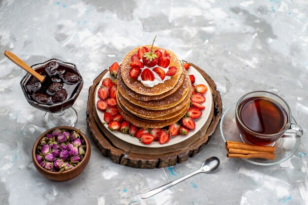 Widok z przodu pyszne okrągłe naleśniki ze śmietaną i czerwonymi truskawkami, herbatą i galaretką na drewnianym biurku tort