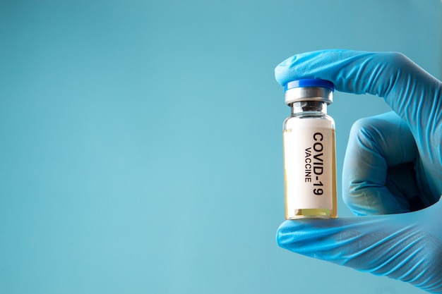 Widok z przodu rękawicy noszącej dłoń trzymającą zamkniętą ampułkę ze szczepionką przeciw covid po lewej stronie na tle niebieskiej fali