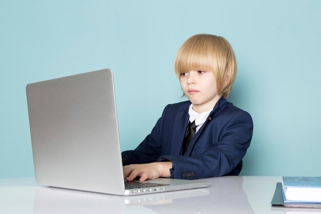 Widok z przodu słodki chłopiec biznesu w niebieskim klasycznym garniturze pozuje przed srebrnym laptopem pracującej mody biznesowej