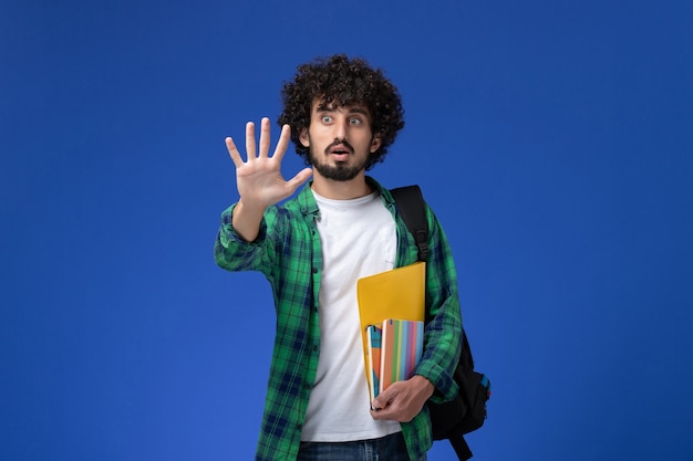 Widok z przodu studenta na sobie czarny plecak, trzymając zeszyty i pliki na niebieskiej ścianie