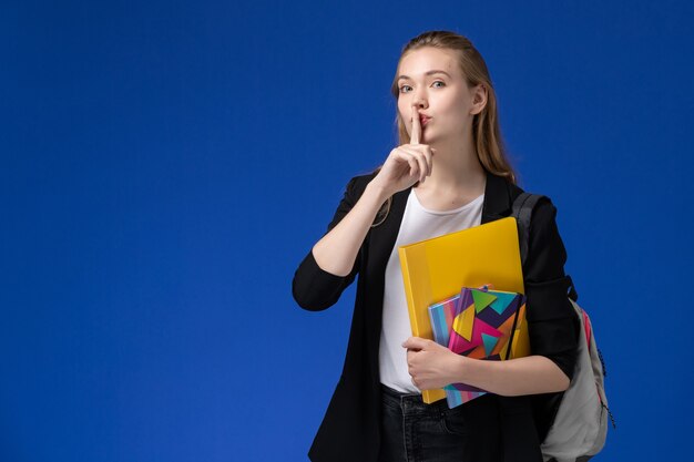 Widok z przodu studentka w białej koszuli i czarnej kurtce na sobie plecak trzymając pliki z zeszytami na niebieskiej ścianie College uniwersyteckie książki lekcyjne