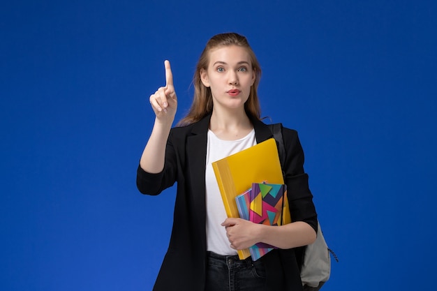 Bezpłatne zdjęcie widok z przodu studentka w czarnej kurtce na sobie plecak trzymając plik z zeszytami na lekcji uniwersytetu na niebieskiej ścianie