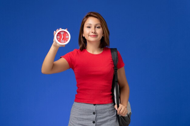 Widok z przodu studentki w czerwonej koszuli z plecakiem trzymając zegary na niebieskiej ścianie