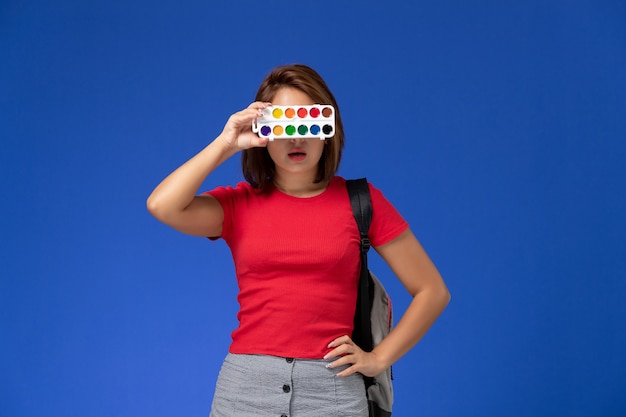 Bezpłatne zdjęcie widok z przodu studentki w czerwonej koszuli z plecakiem trzymającym farby do rysowania na niebieskiej ścianie