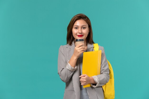 Widok z przodu studentki w szarej kurtce w żółtym plecaku z plikami i kawą na jasnoniebieskiej ścianie