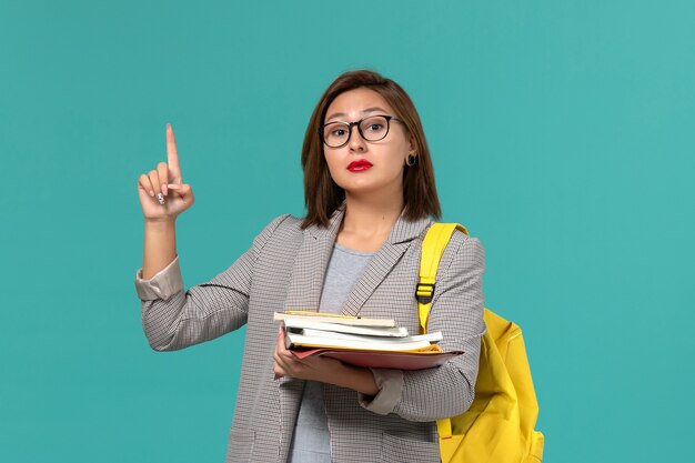 Widok z przodu studentki w szarej kurtce żółtym plecaku trzymając książki na jasnoniebieskiej ścianie
