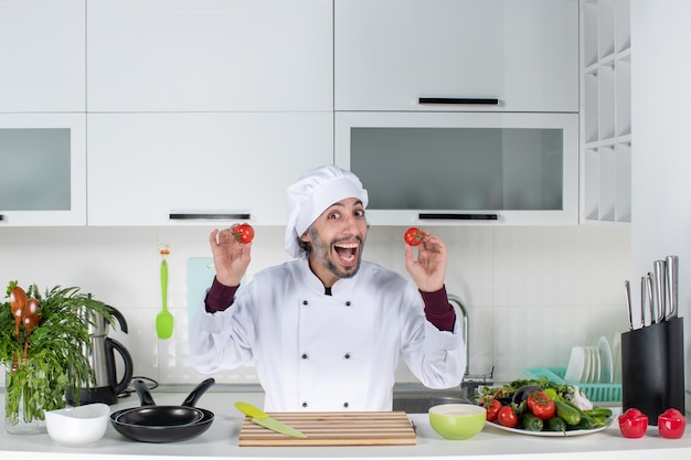 Widok z przodu szczęśliwy męski szef kuchni w mundurze trzymający pomidory w kuchni