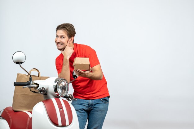 Widok z przodu uśmiechniętego faceta dostawy w czerwonym mundurze stojącego w pobliżu skutera, dając porządek na białym tle